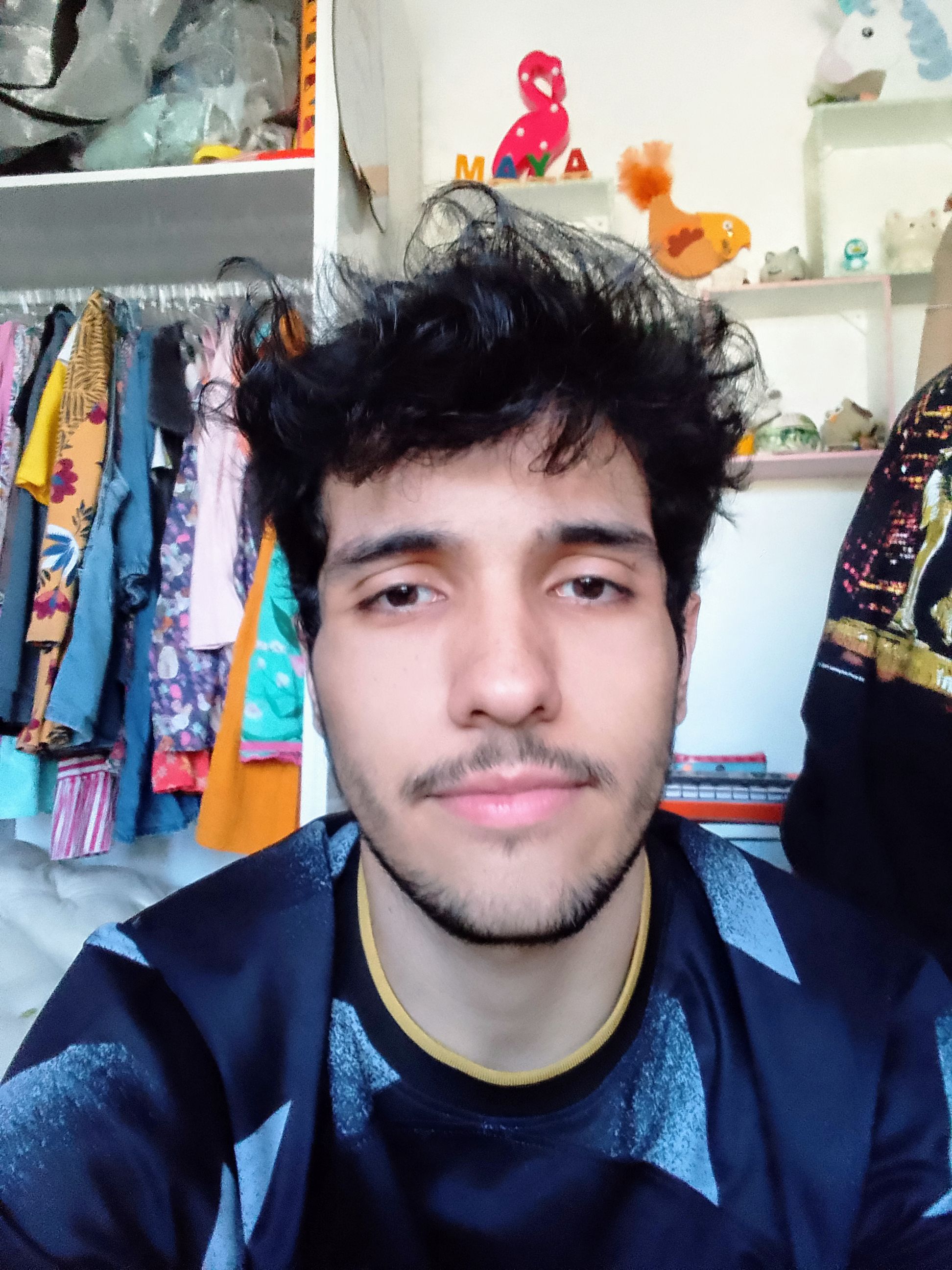 Selfie de Pedro em um quarto, está de camiseta azul com padrões interessantes, uma barba curta e um olhar neutro